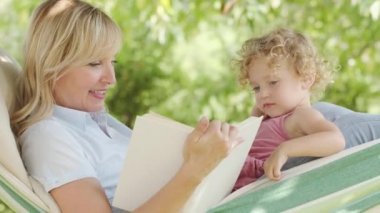 Mutlu anneler günü, kitaptan mavi gözlü küçük kız çocuğuna masal okuyan gülümseyen anneler günü kıvırcık sarı saçlı kız çocuğu, birlikte güneşli bahçede hamakta yatıyor, sevimli aile kavramı.