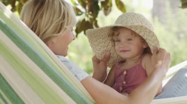 Mutlu anneler günü, gülümseyen anne mavi gözlü küçük kız çocuğuyla oynuyor, hamakta büyük bir hasır şapka takarak onunla oynuyor, bahar ve yaz mevsimi konseptinde, güneşli yeşil bahçede.