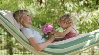 Mutlu anneler günü, gülümseyen anne mavi gözlü küçük kızıyla oynuyor, kız annesine pembe bir buket çiçek veriyor, hamakta güneşli yeşil bahçede yatıyor, bahar ve yaz zamanı konsepti.