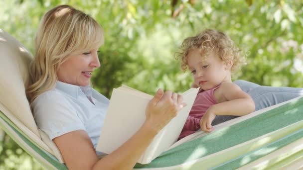 幸せな母の日 笑顔母読書物語から彼女の青い目の小さな女の子の娘子供とともにブロンド髪 一緒に横たわっていますザハンモックで日当たりの良い庭 素敵な家族のコンセプト — ストック動画