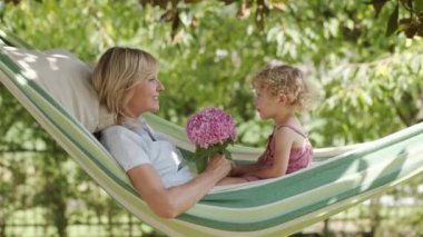 Mutlu anneler günü, gülümseyen anne mavi gözlü küçük kızıyla oynuyor, kız annesine pembe bir buket çiçek veriyor, hamakta güneşli yeşil bahçede yatıyor, bahar ve yaz zamanı konsepti.