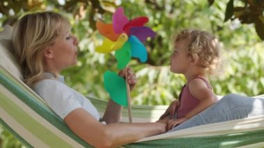 Mutlu gülümseyen anne mavi gözlü küçük kızıyla oynuyor. Sarı kıvırcık saçlı kız çocuğu. Güneşli yeşil bahçedeki hamakta, bahar ve yaz konseptinde, fırıldak içinde birlikte uzanırız.