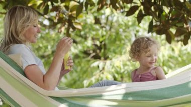 Mutlu anne mavi gözlü küçük kızıyla oynuyor, hamakta birlikte sabun köpüğü üflüyor, yeşil bahçedeki hamakta yatıyor, sarı kıvırcık saçlı kız çocuğu. Bahar ve yaz konsepti