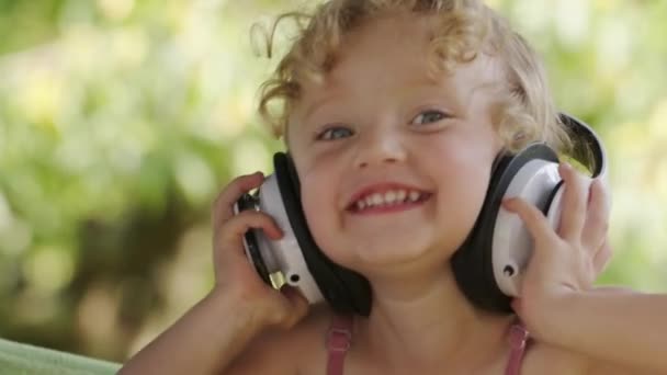 幸せな笑顔の小さな赤ちゃんの女の子は音楽を聞いてヘッドフォンを身に着けています 青い目とブロンド巻き毛を持つ面白い子供のクローズアップポートレート カメラを見て笑う子供 晴れた夏の日に屋外 — ストック動画