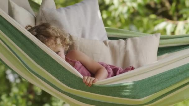 快乐微笑的小女孩挥手打招呼 蓝眼睛卷发的孩子喜欢看镜头 夏天阳光明媚的日子躺在吊床上 在绿色的家花园里 快乐的童年的概念 — 图库视频影像