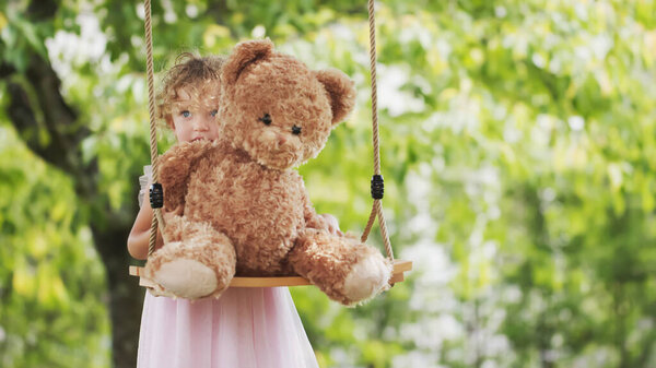 Spring and happy summer time. Joyful smiling little girl playing with teddy bear swinging on the swing. Child with blue eyes and curly blond hair in the green garden at home, concept of healthy growth