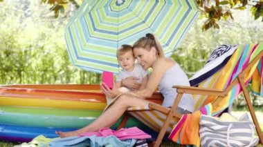 Küçük oğluyla mutlu bir anne, birlikte cep telefonuna bakar ve güneşli bir yaz gününde selfie çekerler, anne iskemlede oturur, çocuk bahçede şişme havuzda oynar.
