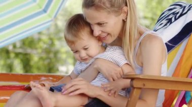 Gülümseyen ve mutlu bir anne bebeğine sarılıyor ve sarılıyor, çocuk güverte sandalyesinde, güneşli bir yaz gününde, ev bahçesinde birlikte oynarken eğleniyor, neşeli bir çocukluk aile hayatı konsepti.