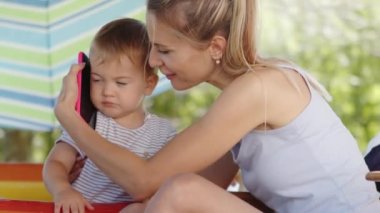 Güneşli bir yaz gününde, küçük oğluyla birlikte cep telefonunda konuşan mutlu bir anne, güverte sandalyesinde oturan bir anne, şişme havuzda oynayan bir çocuk,