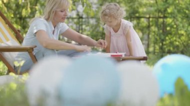 Happy Family on Sunny Summer Day in green garden. Smiling mother on the deck chair playing with her little girl child daughter. Easter holiday, colorful toys and balloons in Backyard Lawn of House.