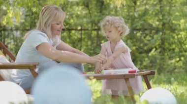 Happy Family on Sunny Summer Day in green garden. Smiling mother on the deck chair playing with her little girl child daughter. Easter holiday, colorful toys and balloons in Backyard Lawn of House.