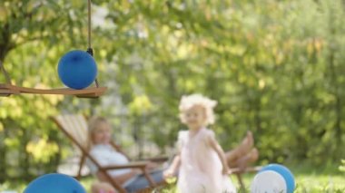 Happy Family on Sunny Summer Day. Smiling mother on the deckchair with laptop computer while her little girl child daughter runs happy in the Backyard Lawn of House, playing with swing and balloons.