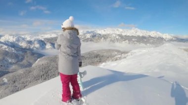 Genç bayan snowboardcu, kışın dağın zirvesinden manzarayı izliyor.