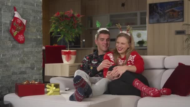 Счастливая пара наслаждается Рождеством на диване, смотря телевизор и обмениваясь подарками — стоковое видео