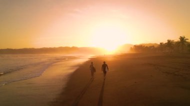 Sörf tahtası ile sahilde yürüyen sörf kankaları. (Sörf tahtası) Sörf arkadaşları gün batımından sonra altın ışıkta gezinip sohbet ediyorlar. Panama 'daki Playa Venao' da güzel bir yaz manzarası.