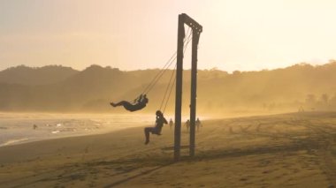 Gün batımında sisli egzotik bir plajda büyük salıncakta sallanan iki arkadaş. Playa Venao 'da salıncakta sallanmak eğlenceli. Tropikal Panama 'da seyahat ederken kaygısız eğlence aktiviteleri.