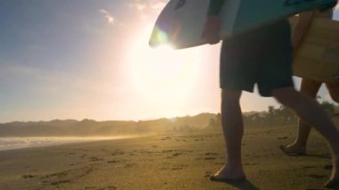 SLOW MOTION, LOW ANGLE VIEW: Couple of surfers walking on a sandy beach with surfboards. Two friends going surfing at sunset on Playa Venao surf spot. Beach lifestyle shot in golden light.