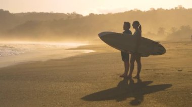 SLOW MOTION: Two surf friends touched by water while standing on the sandy beach and checking waves before surfing. Wave analysis before paddling out for waves. Beach lifestyle shot in golden light.