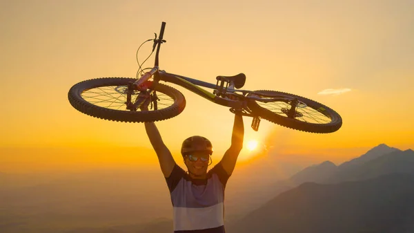 SUN FLARE, CLOSE UP: Happy man lifts his bicycle above his head at sunset after a mountain biking trip in the beautiful Slovenian mountains. Cheerful tourist celebrates winning a mountain bike ride