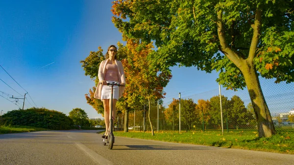 LOW ANGLE: Millennial woman rides an electric scooter down a scenic avenue with gorgeous trees changing colors in autumn. Young Caucasian woman commutes to work on a high tech electric scooter.