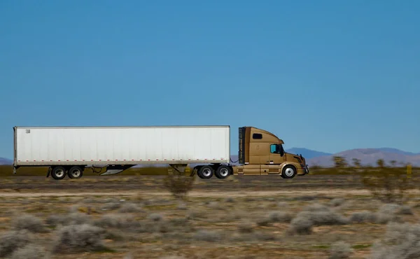 CLOSE UP: Gold colored semi-trailer truck speeds along the interstate highway crossing a desert in United States. Lorry hauls heavy freight container across the countryside of United States of America
