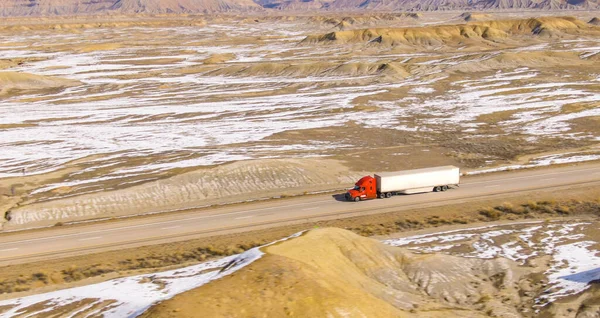 AERIAL: Barren wintry landscape surrounds a cargo truck driving across wintry Utah. Red semi-trailer truck hauls a heavy container across the snowy desert. Truck driving down the interstate freeway.