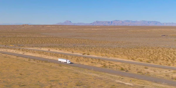 DRONE: Aerial shot of a semi-trailer truck transporting merchandise across the Utah desert on a sunny spring afternoon. White freight truck hauls a heavy cargo container across the barren landscape.