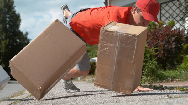 LOW ANGLE, CLOSE UP, DOF: Young male courier trips and falls to the asphalt pavement along with a few parcels. Uncoordinated Caucasian delivery man falls to the ground and drops cardboard boxes.