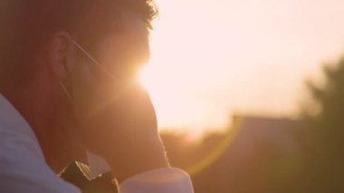 Yavaş Hareket, Yaklaş, El HELD, LENS FLARE, Dof: Genç beyaz adam altın gün batımını izlerken cerrahi maskesini çıkarıyor. İnsan gün doğumunu izlerken maskesini çıkardığı için rahatlar.
