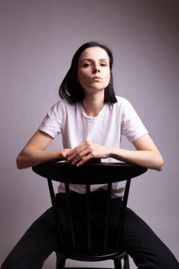 Beyaz tişörtlü kız stüdyoda bir sandalyede oturuyor.