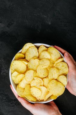 Patates cipsi. Elinde patates cipsi kasesi olan kadın elleri koyu taştan bir arka planda, üst manzarada fotokopi alanı var..