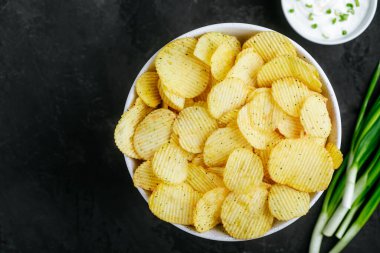 Patates cipsi. Çıtır patates cipsi ve soğanlı ekşi krema. Koyu taştan bir arka planda, fotokopi alanı olan üst manzara..