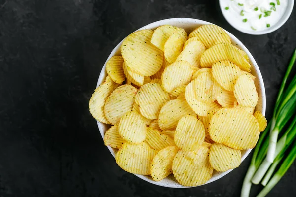 Patates cipsi. Çıtır patates cipsi ve soğanlı ekşi krema. Koyu taştan bir arka planda, fotokopi alanı olan üst manzara..