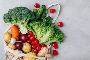 Sebzeli ağ torbası. Organik brokoli, domates, salatalık, havuç, soğan, limon, sarımsak, zencefil, marul ve patates gri taştan arka planda. Sıfır atık, plastiksiz konsept. Sağlıklı beslenme ve detoks. Üst görünüm w
