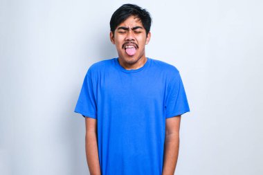 Beyaz arka planda dikilen sıradan gömlekli yakışıklı Asyalı çocuk komik bir ifadeyle dilini dışarı çıkartıyor. Duygu kavramı.