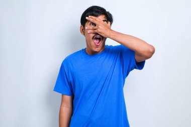 Soyutlanmış beyaz arka planda genç yakışıklı bir adam. Şoka girmiş, yüzünü ve gözlerini kapamış, utangaç ifadesiyle parmaklarının arasından bakıyor..