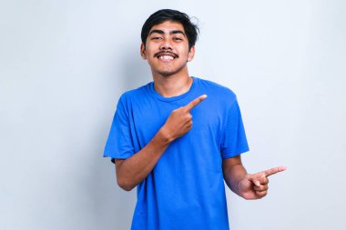 Genç yakışıklı Asyalı bir adam beyaz arka plan üzerine gündelik gömlek giyiyor, yüzünde büyük bir gülümseme ile; kameraya el parmağı ile bakıyor.
