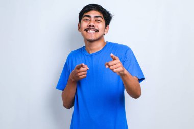 Beyaz arka plan üzerinde duran Asyalı genç ve yakışıklı bir adam parmaklarıyla seni ve kamerayı işaret ediyor, neşeli ve pozitif gülümsüyor.