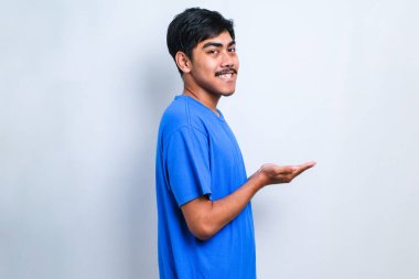 İzole edilmiş beyaz arka plan üzerine gündelik tişört giyen genç yakışıklı adam avuç içleri açık, fotokopi alanını gösteren, gülümseyerek mutlu bir gülümseme sunan,