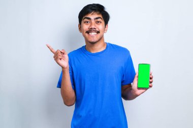 Mavi tişörtlü mutlu Asyalı adam kameraya gülümserken elinde akıllı telefonuyla beyaz arkaplanı işaret ediyor.
