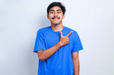 Genç yakışıklı Asyalı bir adam beyaz arka plan üzerine gündelik gömlek giyiyor, yüzünde büyük bir gülümseme ile; kameraya el parmağı ile bakıyor.