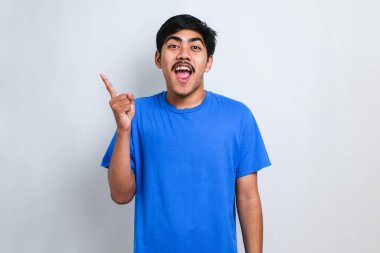 Genç yakışıklı Asyalı bir adam beyaz arka plan üzerine gündelik gömlek giyiyor, yüzünde büyük bir gülümseme ile; kameraya el parmağı ile bakıyor.