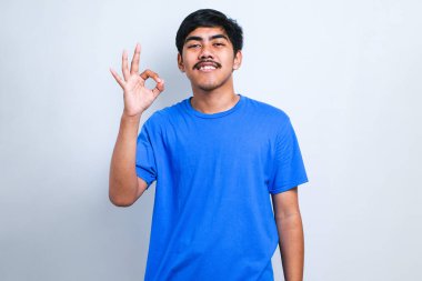 Bıyıklı yakışıklı genç adam beyaz arka plan üzerine günlük tişört giyip gülümsüyor ve el ve parmaklarıyla tamam işareti yapıyor. başarılı ifade.