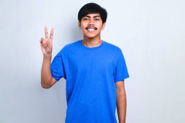 Beyaz arka planda sıradan tişörtler giyen yakışıklı Asyalı genç. Barış sembolü yapıyor. Yüzünde parmaklar, gülümseyerek zaferi gösteriyor.
