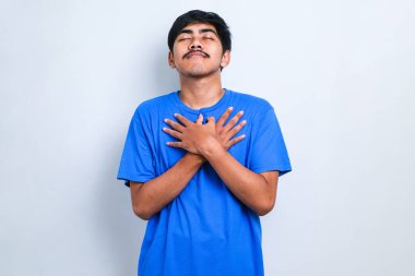 Yakışıklı, sıradan tişörtler giyen, elleri göğsünde, gözleri kapalı gülümseyen ve beyaz arka planda minnet dolu bir jest yapan genç adam. sağlık kavramı.