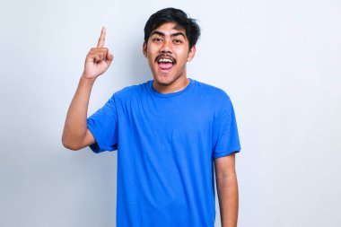 Beyaz arka plan üzerine gündelik tişört giyen genç yakışıklı adam başarılı bir fikirle işaret ediyor. Heyecanlı ve mutlu. Bir numara..
