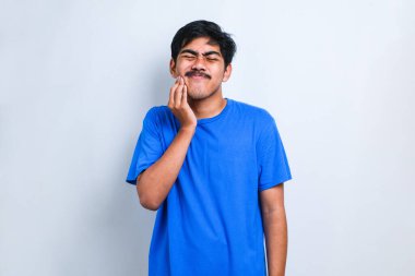 İzole edilmiş beyaz arka plan üzerinde duran sıradan tişört giyen genç yakışıklı diş ağrısı ya da diş ağrısı yüzünden ağız ve acı dolu yüz ifadelerine dokunuyor. Dişçi