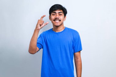 Beyaz arka planda sıradan tişörtler giyen genç ve yakışıklı bir adam gülümsüyor ve el hareketleriyle küçük beden işareti yaparak kameraya bakıyor. ölçüm kavramı.