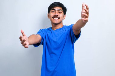 Genç yakışıklı Asyalı sıradan bir tişört giymiş kameraya bakıp kucaklaşmak için gülümsüyor. Beyaz arka planda mutluluğu kucaklayan neşeli ifade