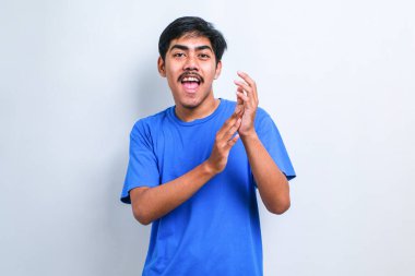 Beyaz arka plan üzerine gündelik tişört giyen genç yakışıklı adam alkışlıyor ve mutlu, neşeli ve gururlu elleri bir arada alkışlıyor.